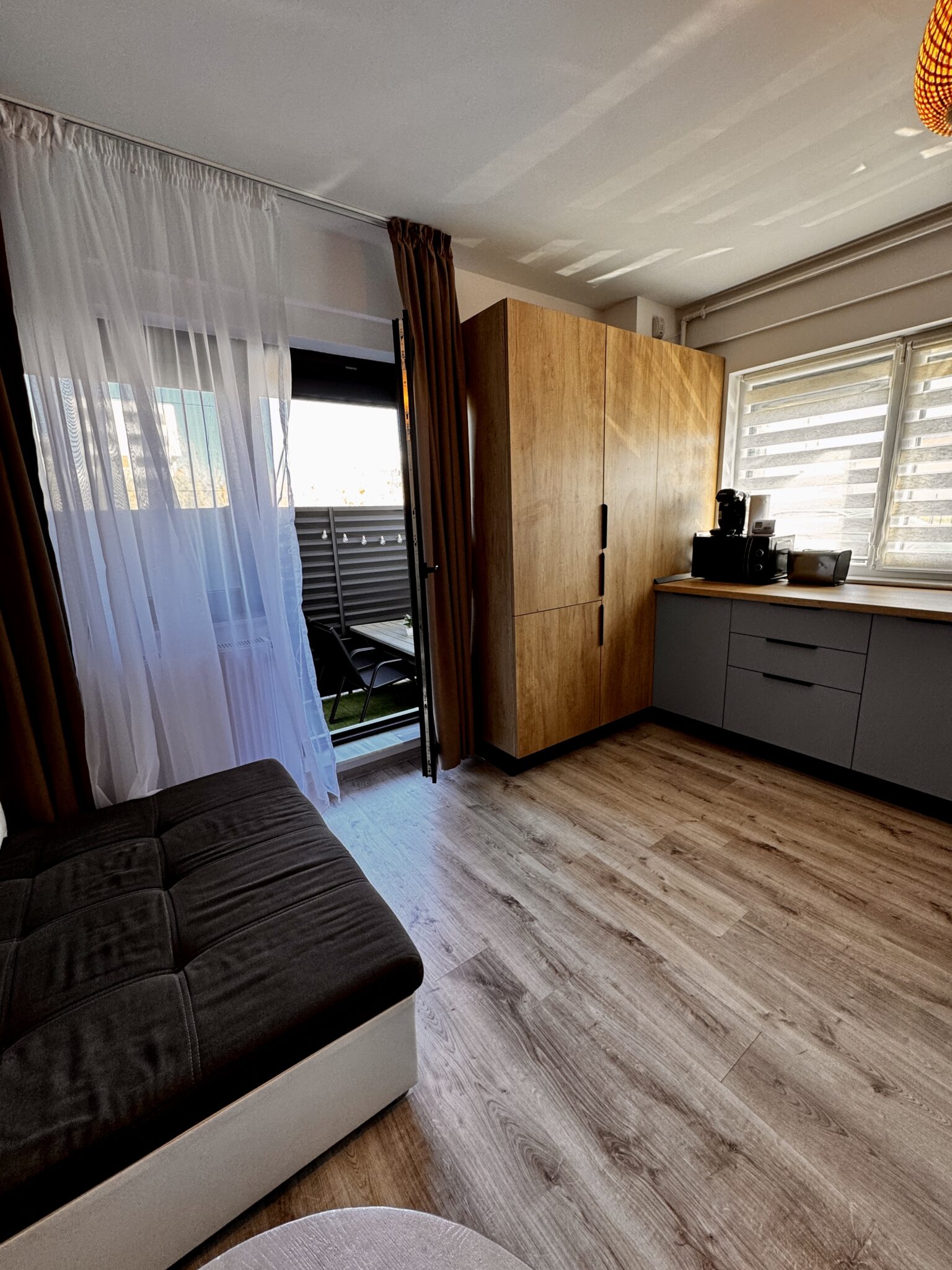 Cameră modernă în Mamaia Nord, cu parchet, o canapea maro și albă, un dulap mare, jaluzele la fereastră, un aparat de cafea pe blat și o ușă care duce spre un mic balcon – descoperiți confortul în apartamentul 2 camere de la B2 Home15.