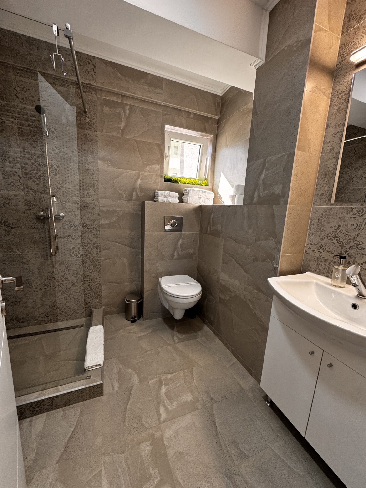 Baie modernă în Apartamentul cu 2 camere, B2 Home15, Mamaia Nord, cu gresie gri, cabină de duș din sticlă, toaletă montată pe perete cu o fereastră mică deasupra și un dulap alb pentru chiuvetă cu oglindă.