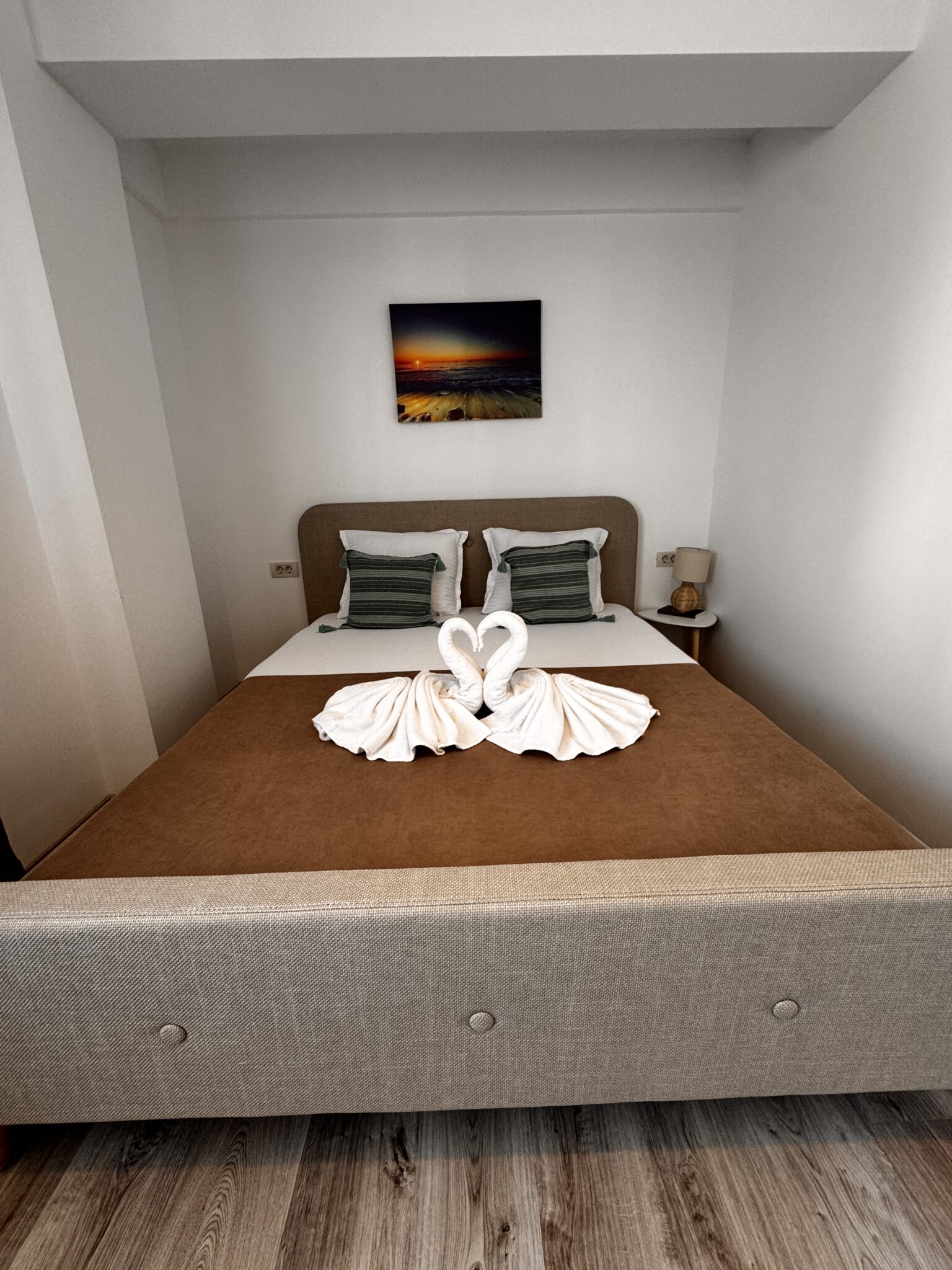 Un pat îngrijit în camera Apartament 2 de la B2 Home15, cu lebede din prosoape care formează o inimă, perne decorative verzi, o pătură maro și o fotografie înrămată cu un apus deasupra tăbliei patului din Mamaia Nord.