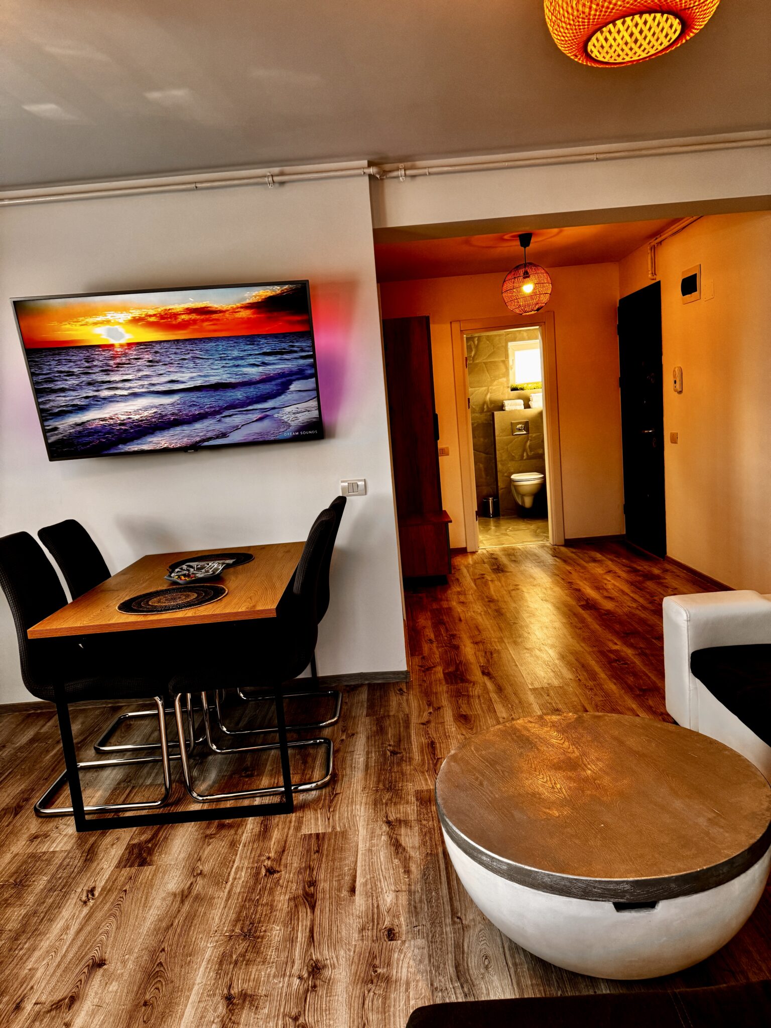 Interior modern al unui apartament de la B2 Home15 din Mamaia Nord, cu un televizor montat pe perete care afișează un apus de soare, masă cu scaune, parchet și vedere spre baie – perfect pentru relaxare în acest apartament cu 2 camere.