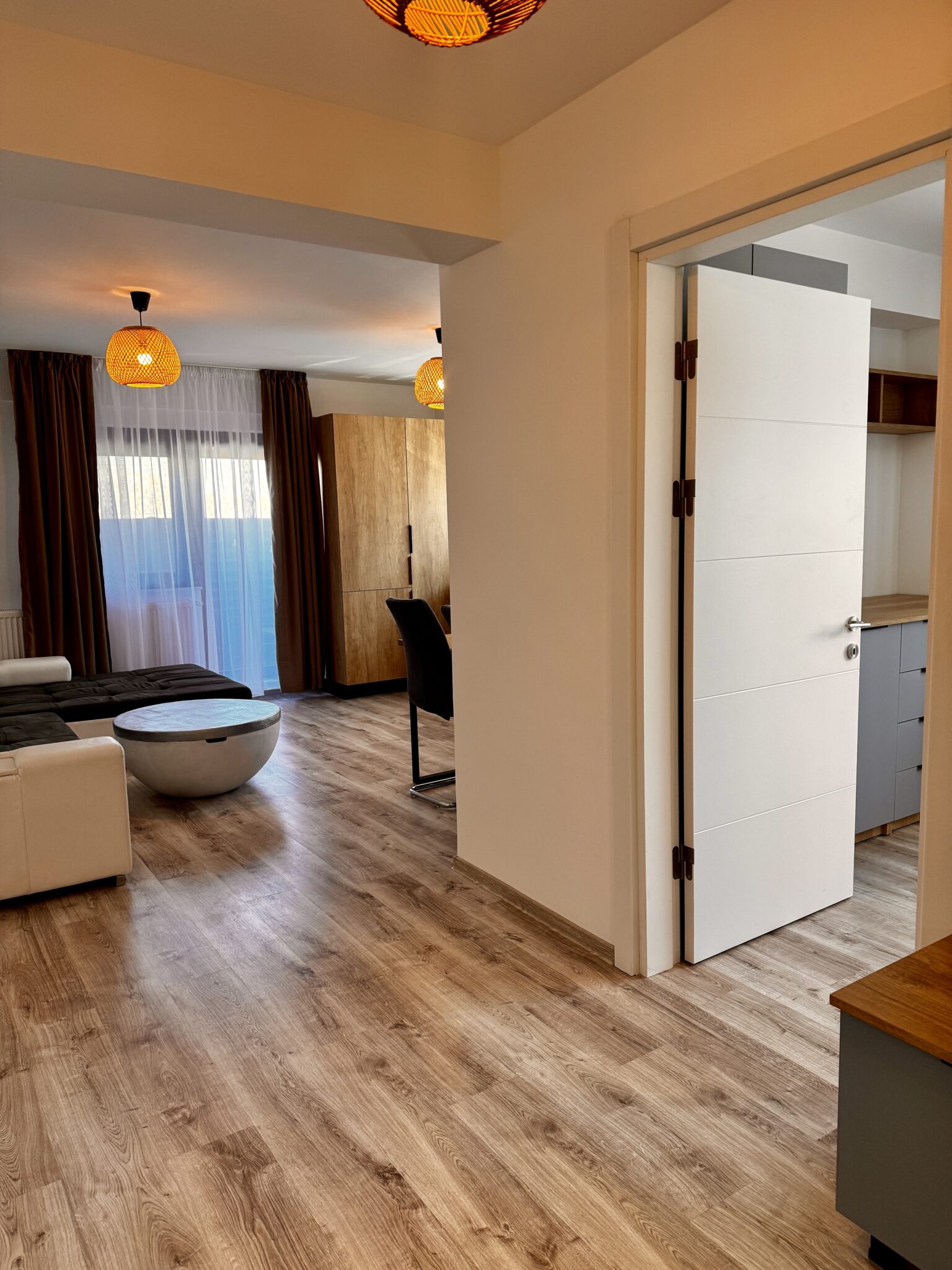 Descoperiți B2 Home15 în Mamaia Nord — o garsonieră modernă și ordonată, cu parchet, un pat, o canapea, o măsuță de cafea rotundă, un birou și o fereastră mare, împodobită cu perdele transparente și maro.