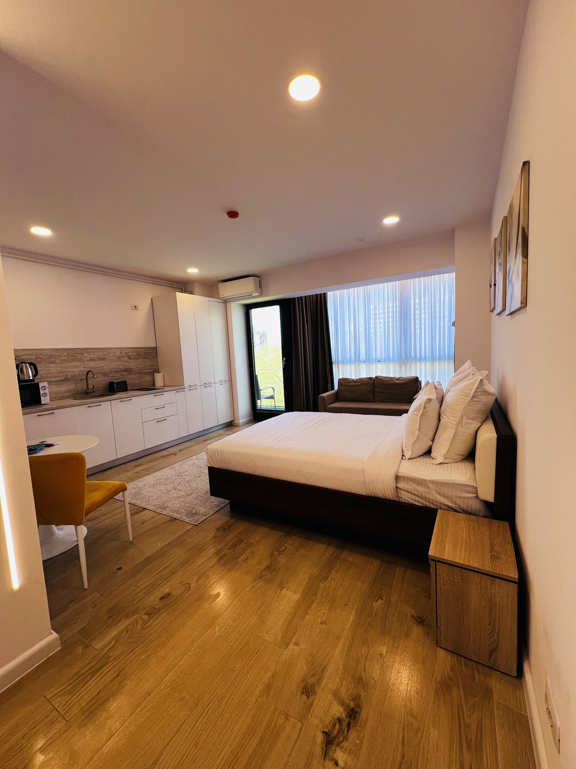 Apartament studio modern în Mamaia, cu pat, canapea, chicinetă, masă și fereastră mare cu perdele, parchet și decor minimalist.