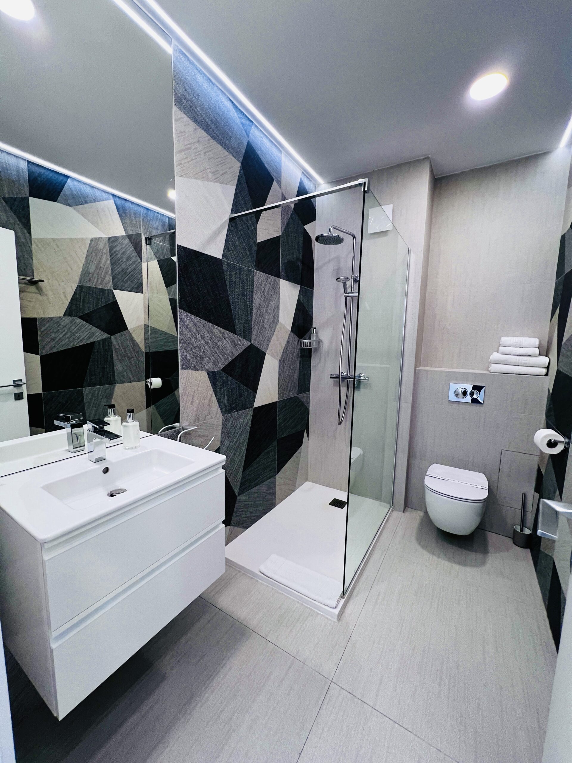 Baie modernă într-un studio din Mamaia, cu un perete geometric decorativ, o cabină de duș din sticlă, o toaletă montată pe perete, o chiuvetă albă și prosoape împăturite cu grijă pe un raft.