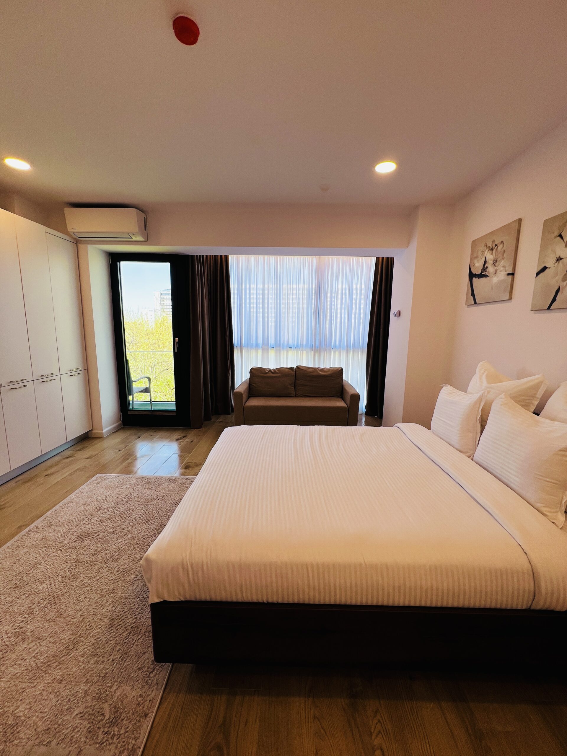 Un studio modern în Mamaia, cu un pat mare cu lenjerie albă, o canapea confortabilă lângă fereastră, dulapuri elegante încorporate și o ușă care duce spre un balcon privat.