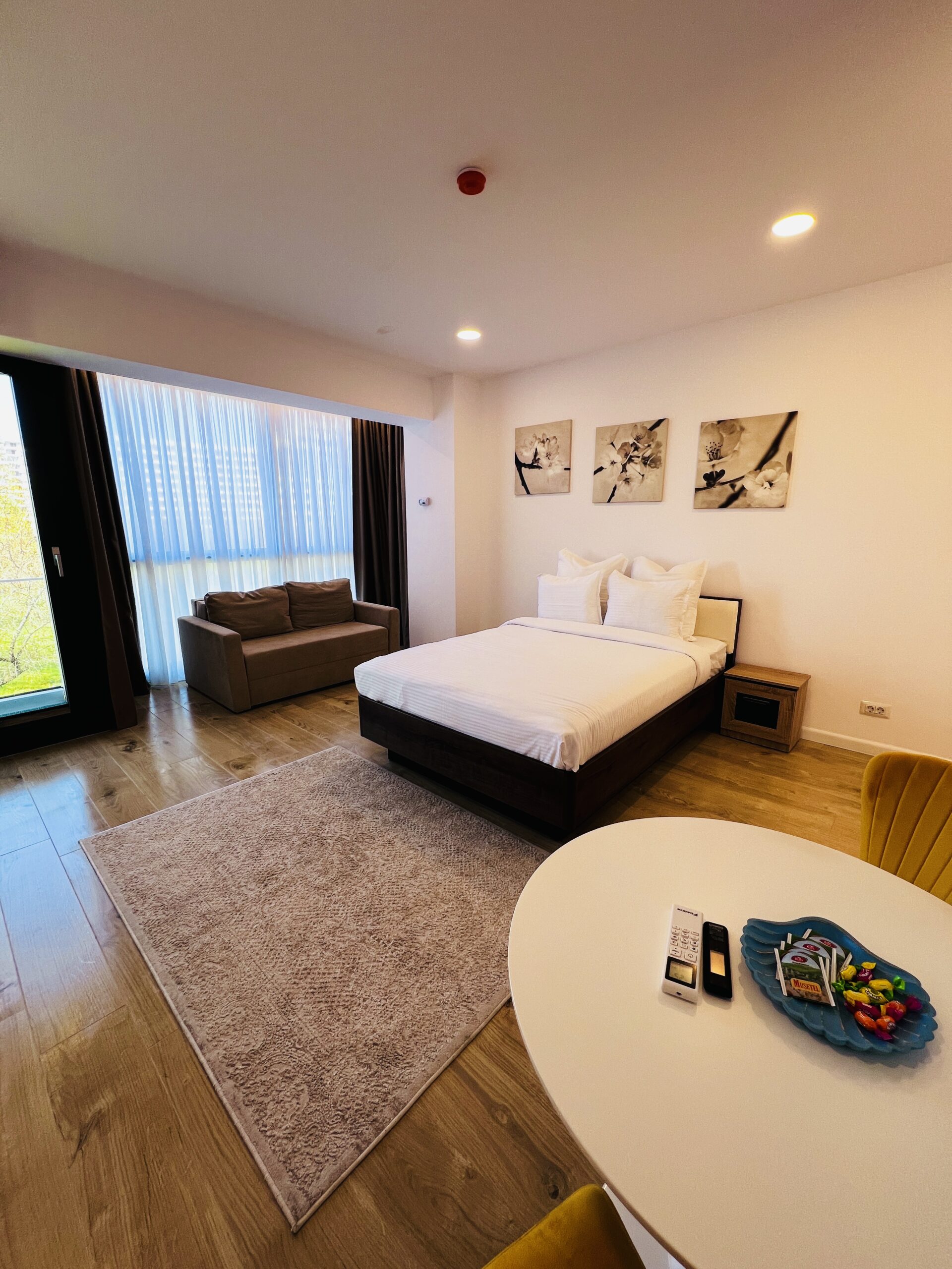 O garsonieră modernă în Mamaia, cu un pat îngrijit, o canapea gri lângă ferestre mari, o noptieră mică, opere de artă elegante deasupra patului, o masă rotundă și un scaun galben vibrant.