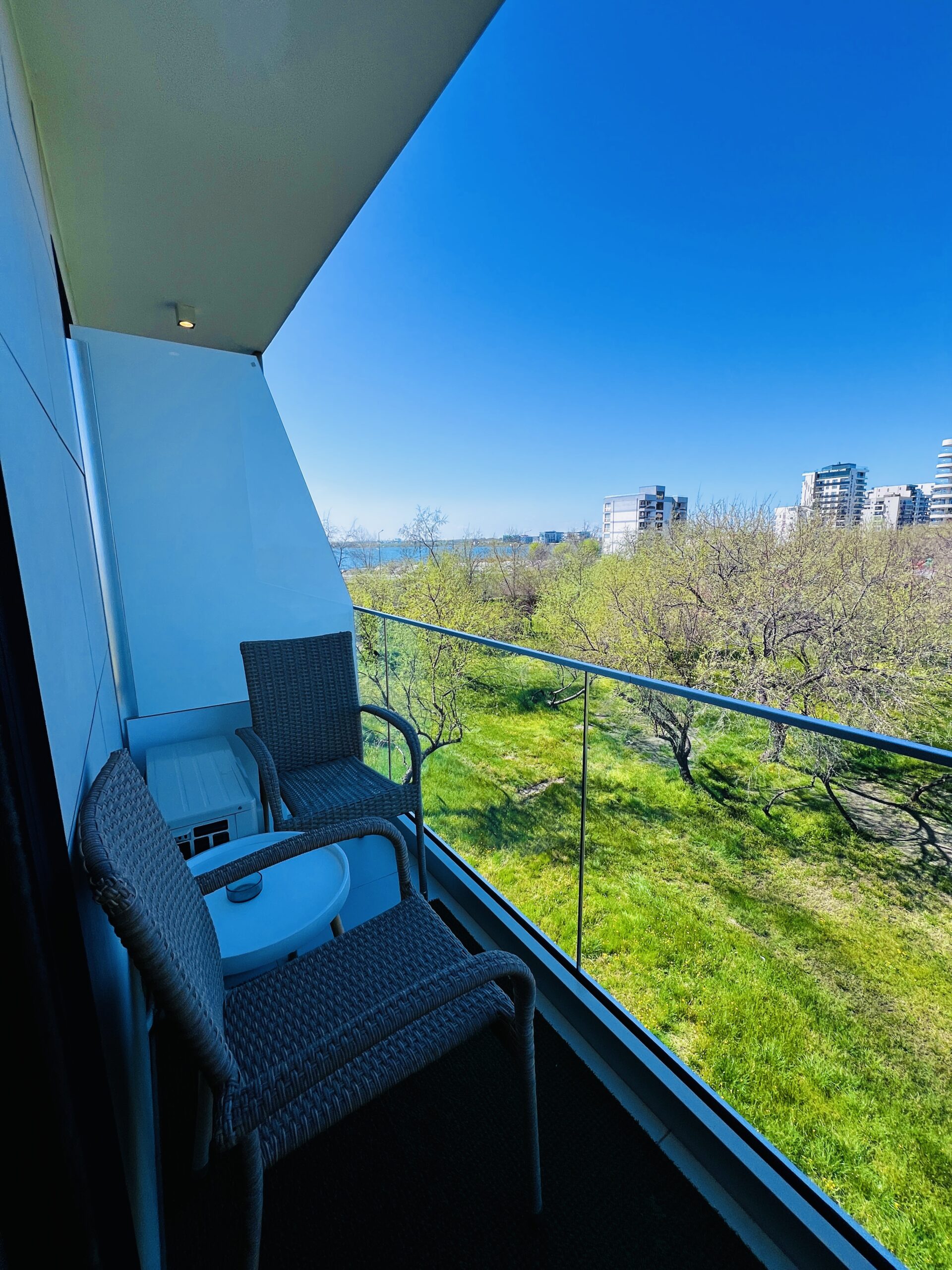 Un mic balcon de la această garsonieră din Mamaia are două scaune din răchită și o masă, cu vedere la o zonă cu iarbă și la clădiri moderne îndepărtate sub un cer albastru senin.