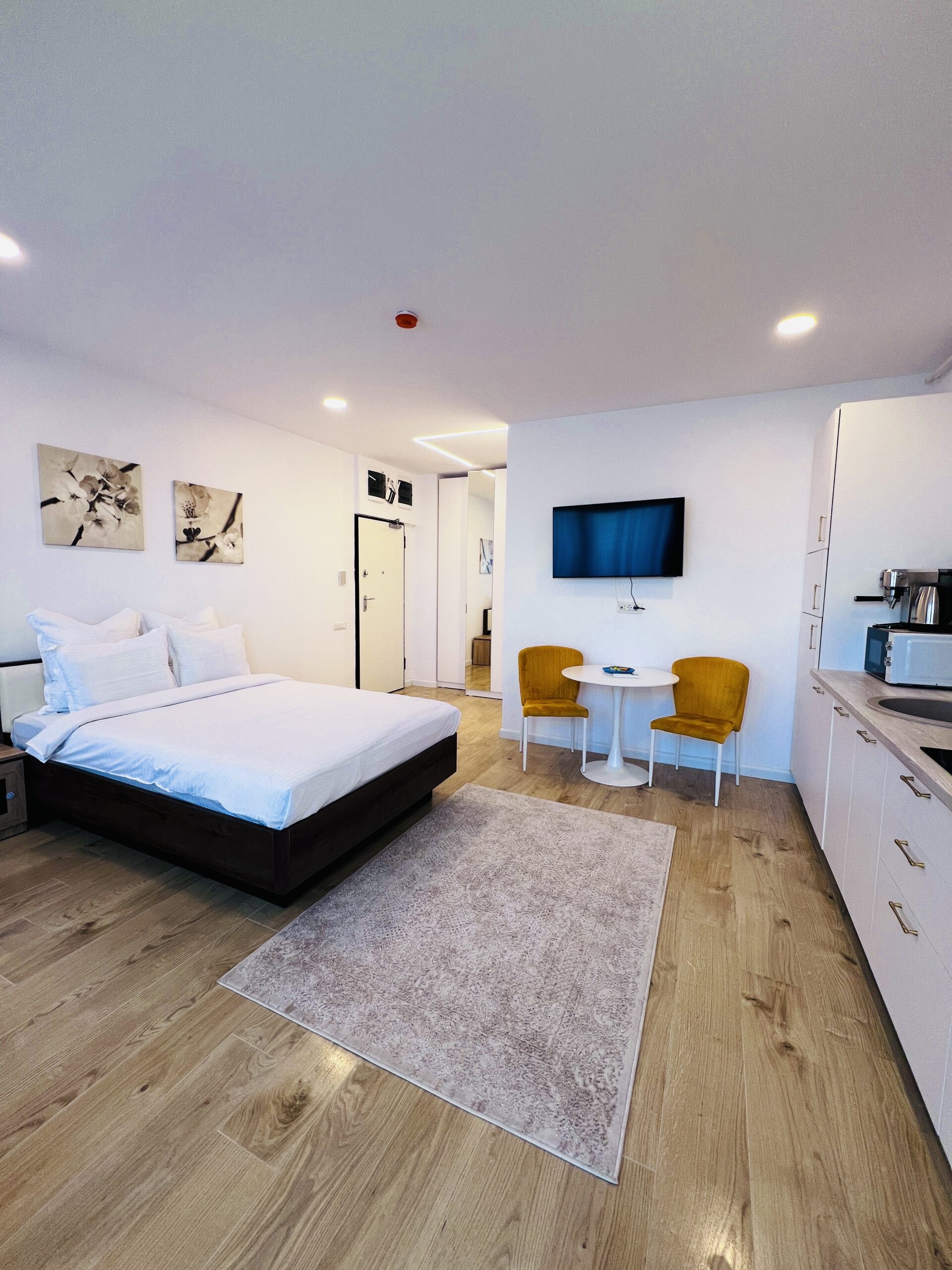 Studio modern în Mamaia, cu pat dublu, televizor montat pe perete, masă mică cu două scaune galbene, chicinetă și parchet din lemn deschis la culoare.