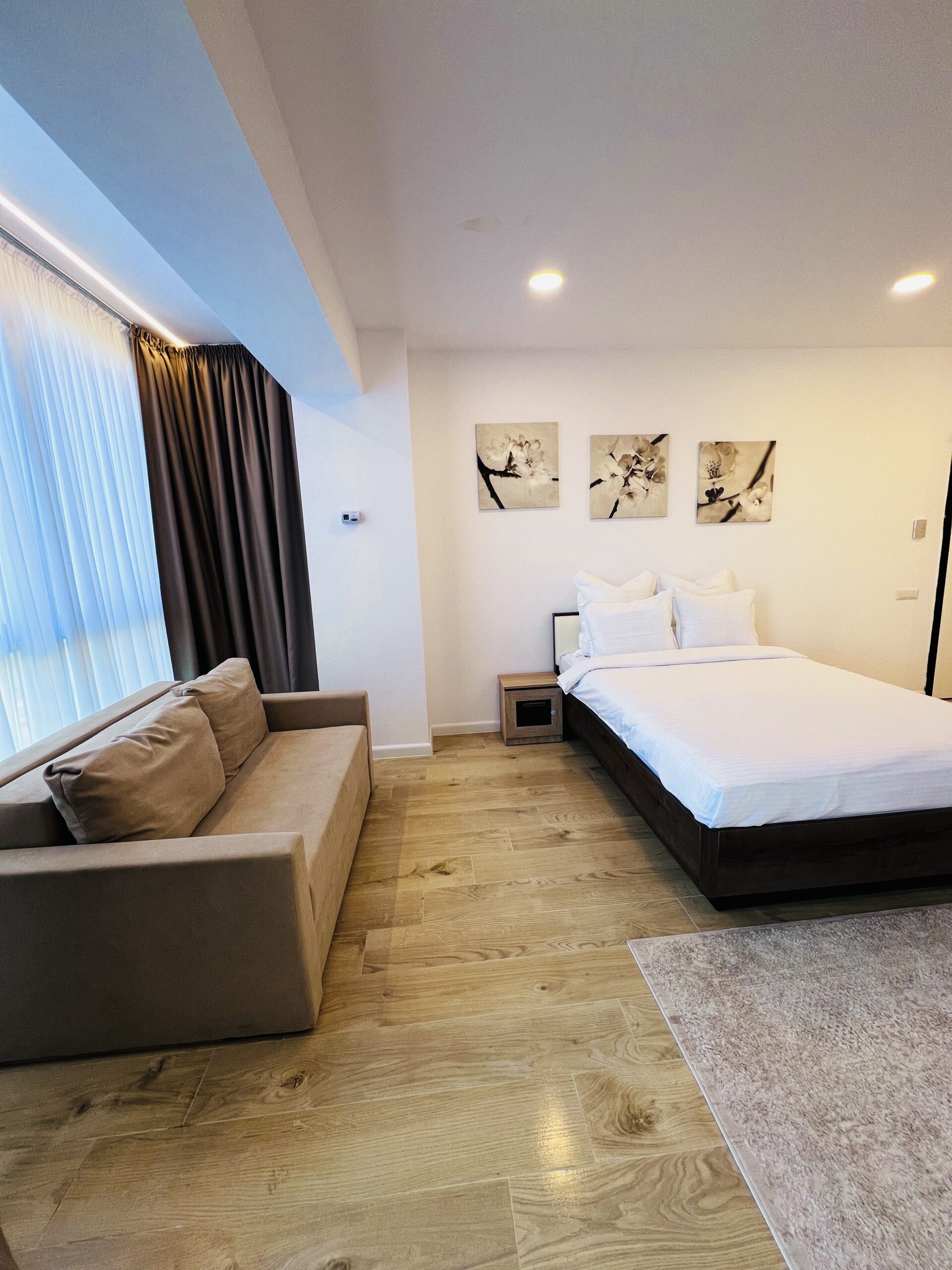 Un studio modern în Mamaia, cu parchet din lemn deschis la culoare, o canapea bej, un pat îngrijit, trei opere de artă deasupra patului și ferestre mari cu perdele transparente.