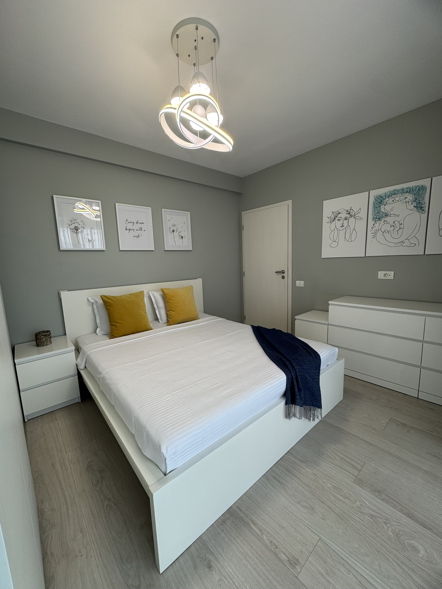 Dormitor modern într-un apartament cu 3 camere în Mamaia, cu pat alb cu perne galbene, pătură albastră, mobilier alb, decor minimalist și tablouri înrămate pe pereți gri, sub o lampă de tavan contemporană.