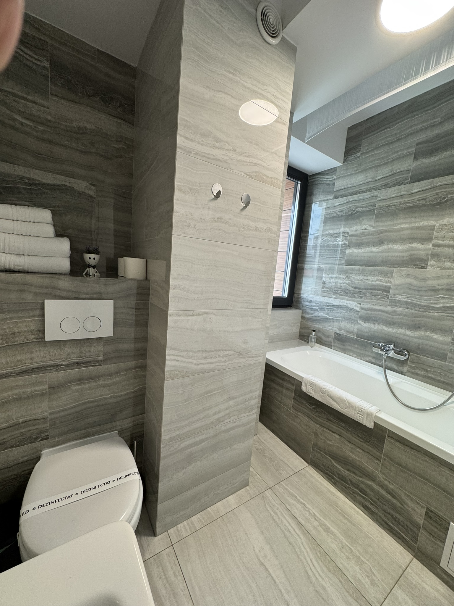 Baie modernă într-un apartament cu 3 camere din Mamaia, cu gresie din marmură gri, toaletă montată pe perete, prosoape suprapuse, cadă cu duș portabil și o fereastră care oferă lumină naturală.