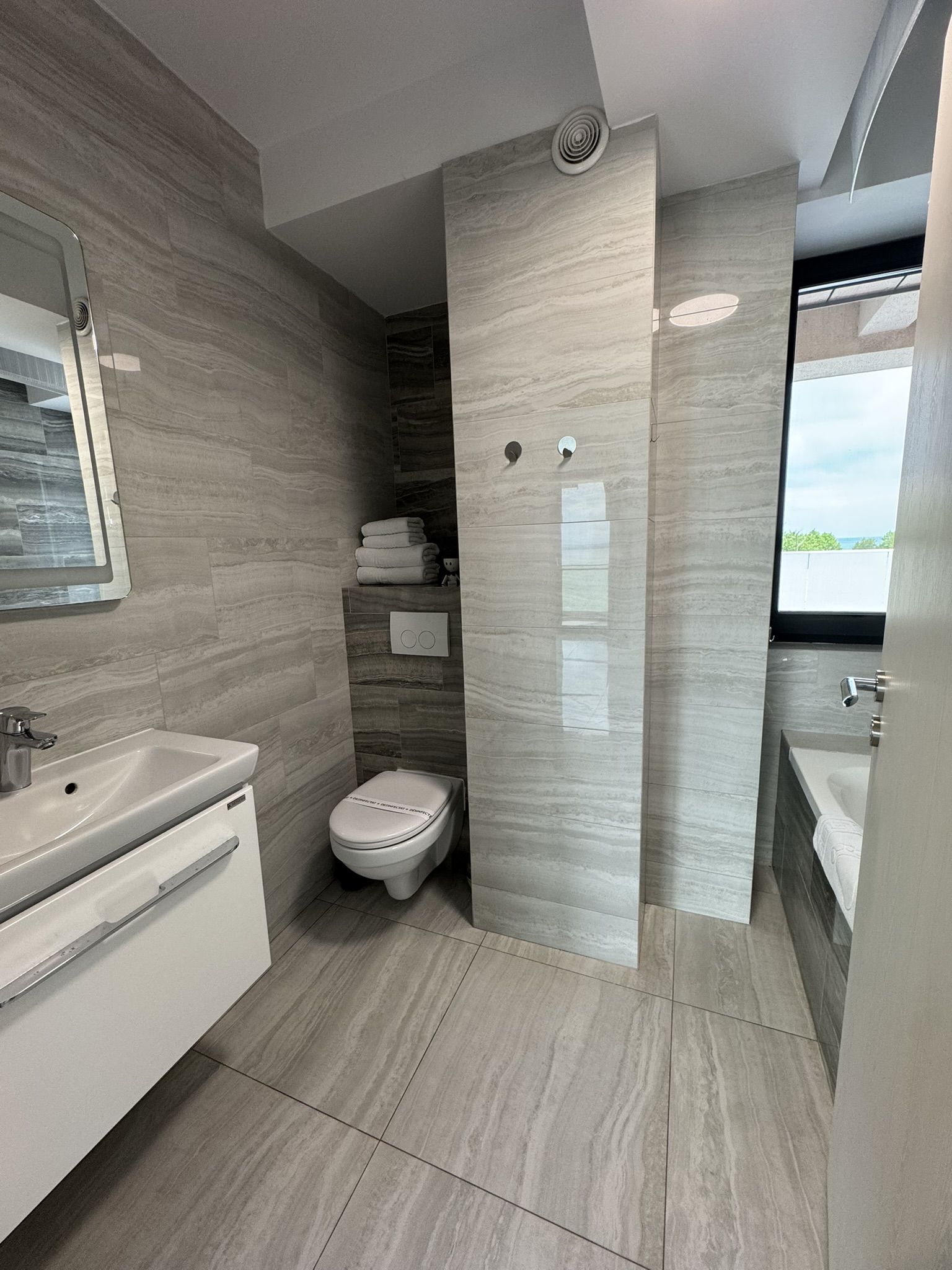 Baie modernă într-un apartament cu 3 camere din Mamaia, cu pereți și podele din gresie gri, chiuvetă și toaletă montate pe perete, prosoape suprapuse, un separator parțial și o fereastră lângă cadă.