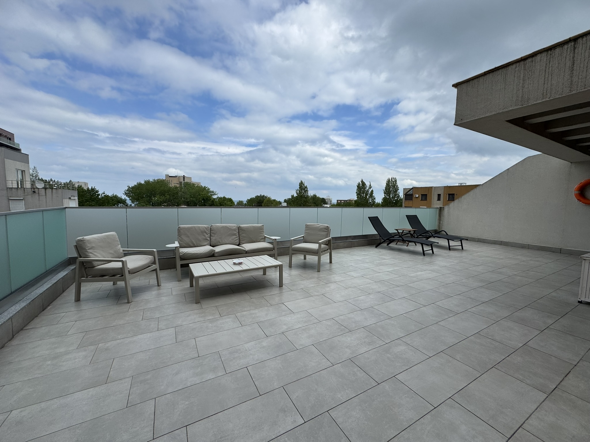 Terasă exterioară spațioasă, cu pardoseală cu gresie, o canapea bej așezată în jurul unei măsuțe de cafea, două șezlonguri negre și o balustradă de sticlă – ideală pentru relaxare în timpul sejurului dumneavoastră în apartamentul cu 3 camere Cazare Mamaia, sub cerul parțial înnorat.