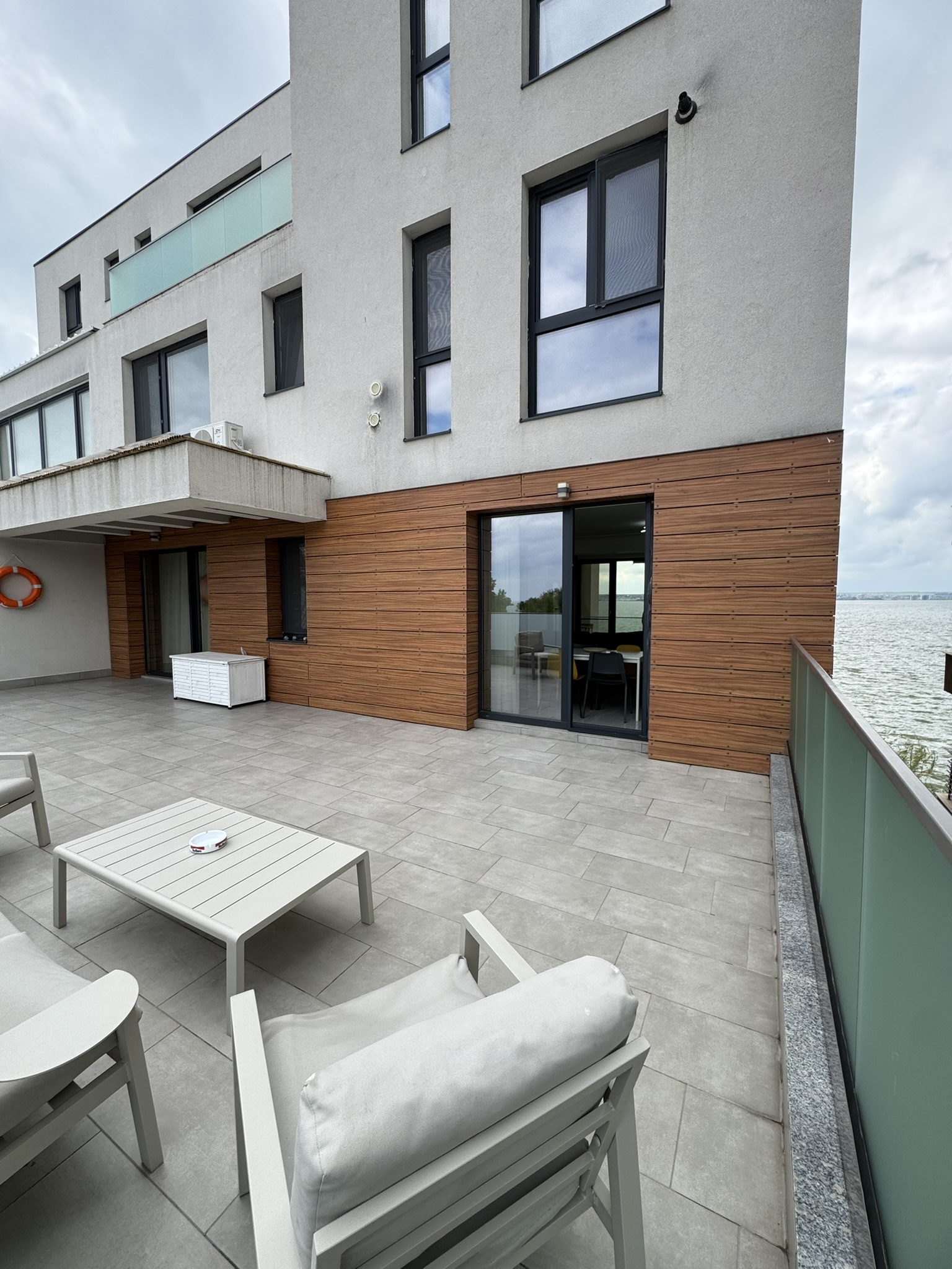 Apartament modern în Mamaia, cu 3 camere, cu ferestre mari și lambriuri din lemn, precum și o terasă spațioasă cu gresie și mobilier de exterior, cu vedere la o întindere de apă.