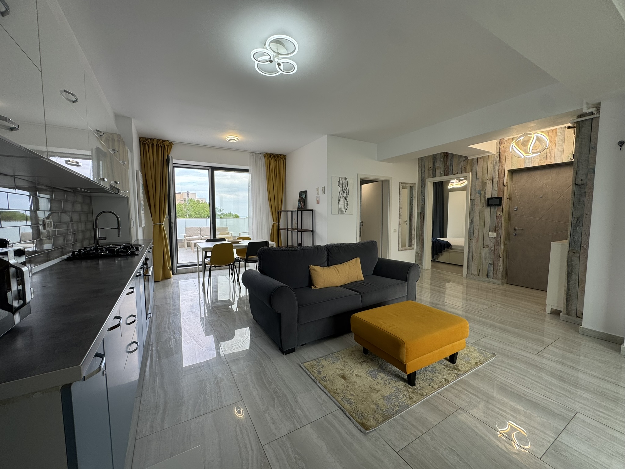 Acest apartament cu 3 camere din Mamaia are un living modern, open space, cu o bucătărie elegantă, o canapea, un otoman galben, o masă, ferestre mari cu perdele galbene și un hol care duce spre alte camere.