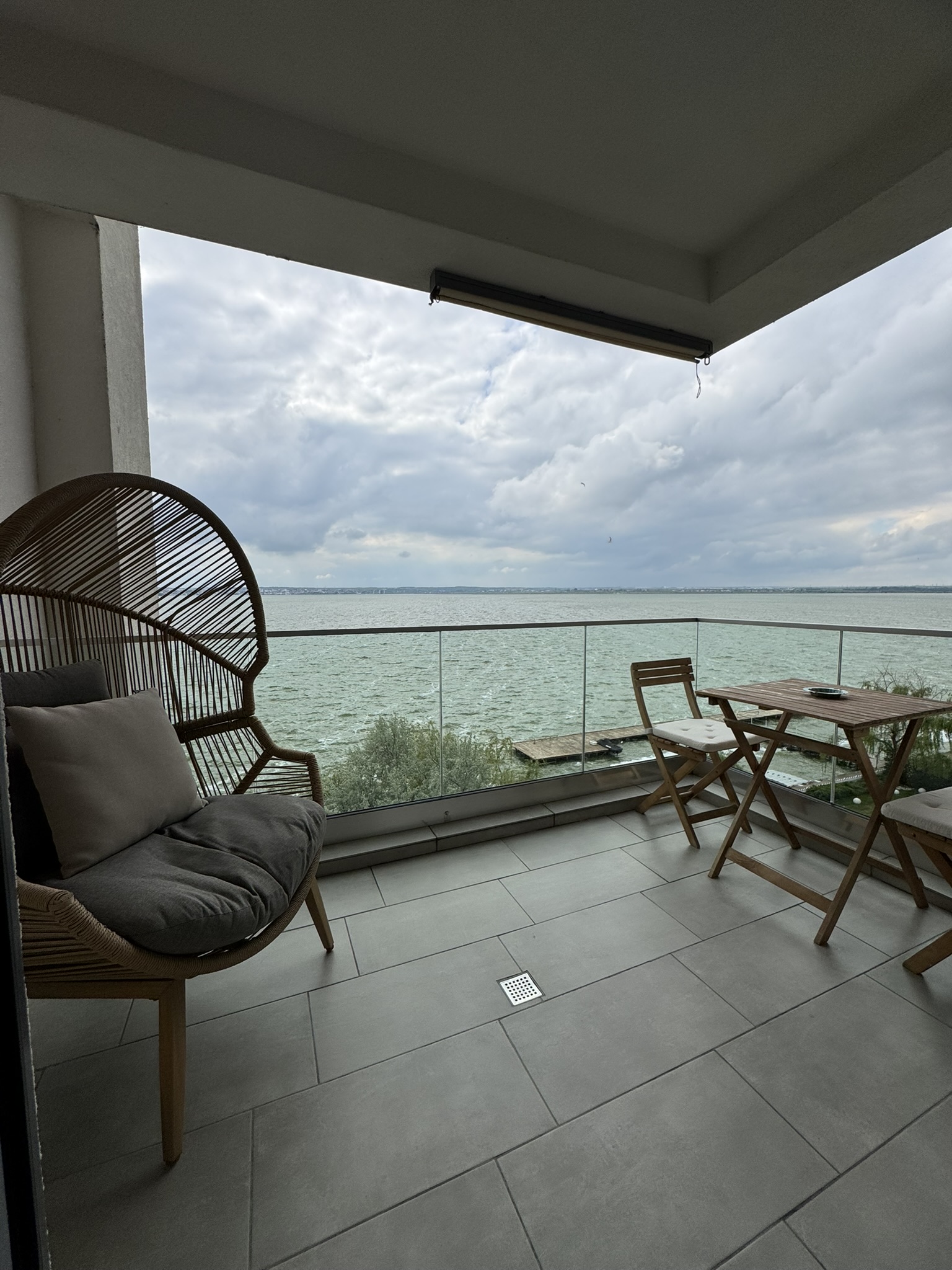 Acest apartament cu 3 camere din Mamaia are un balcon acoperit, un scaun din răchită, o masă mică din lemn și scaune, podea cu gresie și o balustradă de sticlă cu vedere la cerul înnorat și la marea calmă.